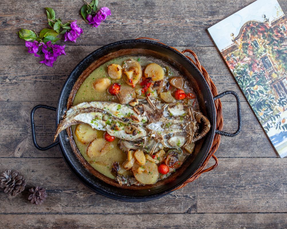 A rustic fish and potato dish at Can Travi Nou, served on a round plate beside flowers and artwork