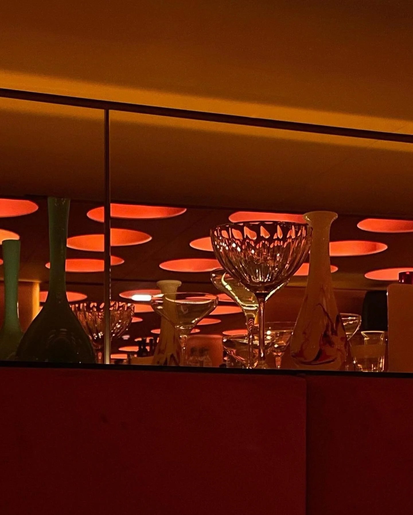 Vintage coupes and glass vases on a mirrored ledge reflecting the orange ceiling lights at Naked & Famous