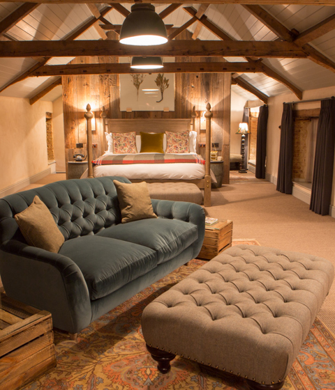 A beamed bedroom at The Pig at Combe with a velvet sofa, tufted ottoman and four-poster bed