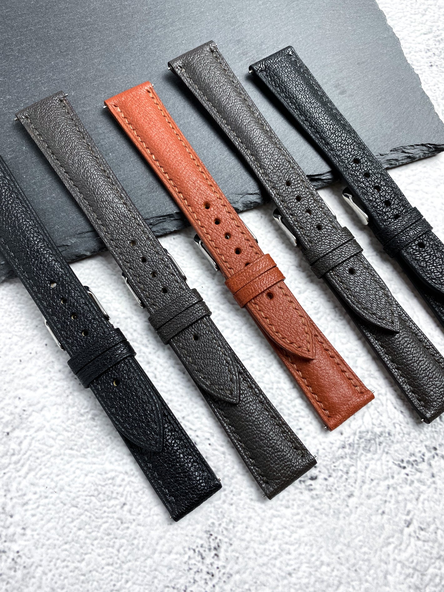 Five handmade leather watch straps in black, grey and tan arranged on stone