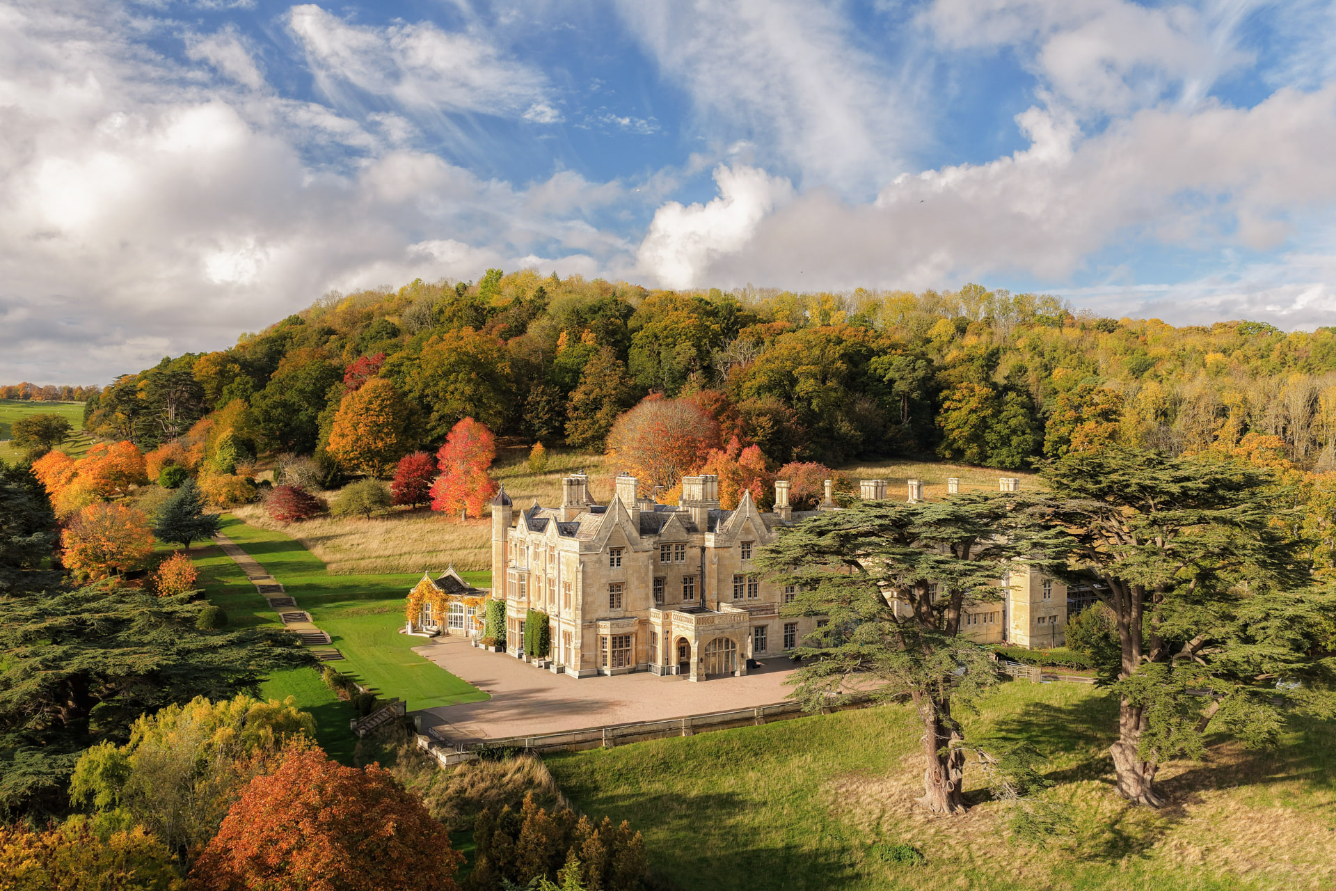 Dumbleton Hall from the air — the golden-stone Tudor Revival house set against its wooded hillside in autumn