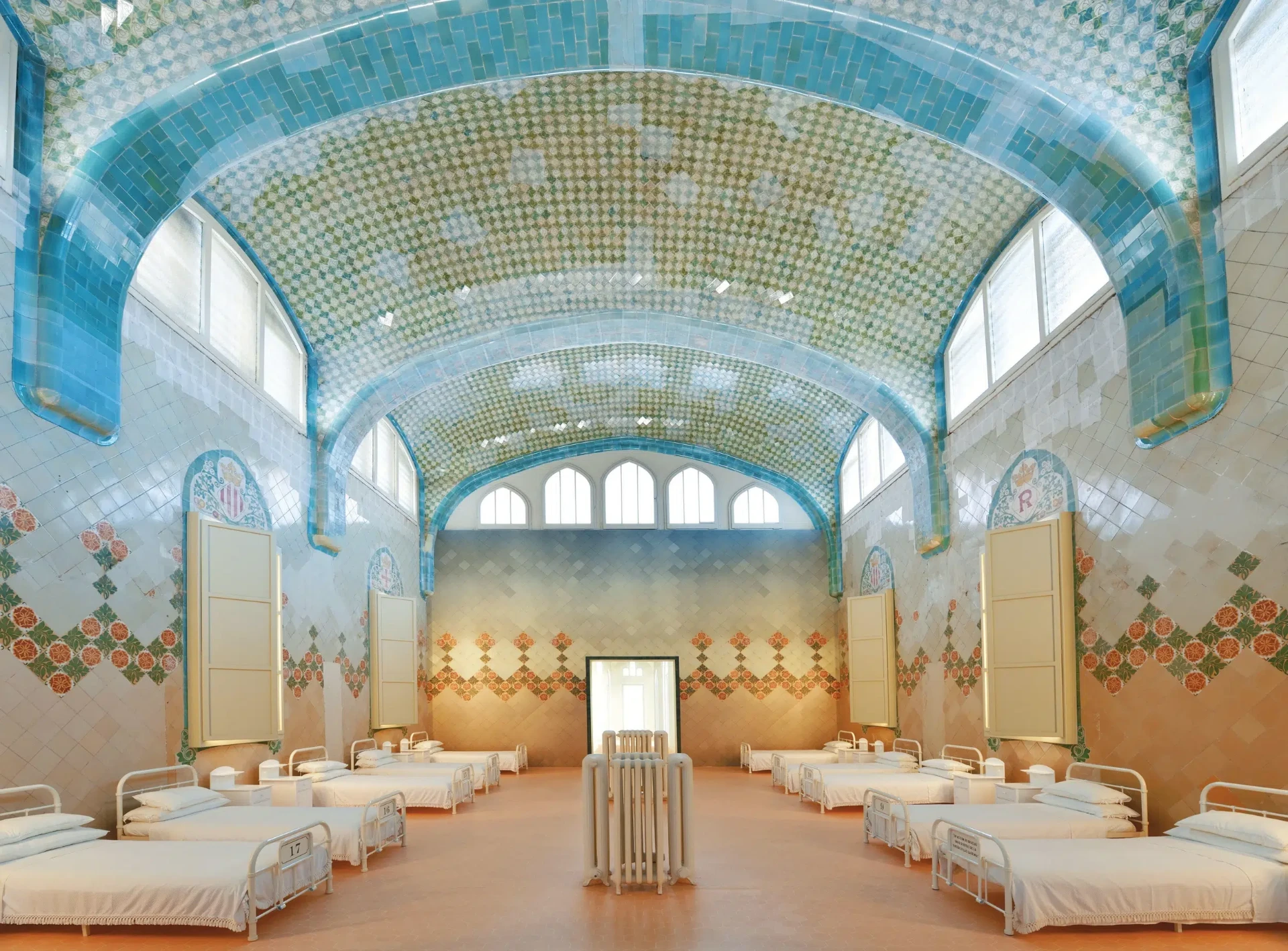A restored Sant Pau hospital ward with white beds, tiled walls, floral ceramics and blue arched ceilings