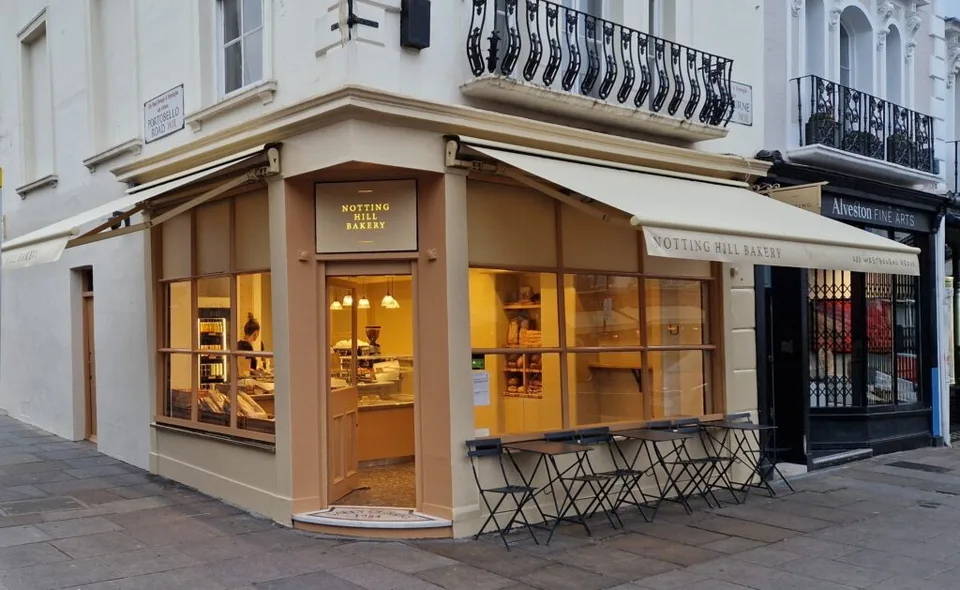 The corner shopfront of Notting Hill Bakery on Westbourne Grove, with cream awnings and café tables outside