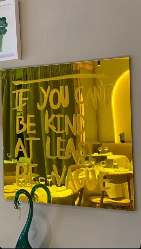 A gold mirror painted with the words “If you can’t be kind, at least be vague”, with a dining room reflected in it