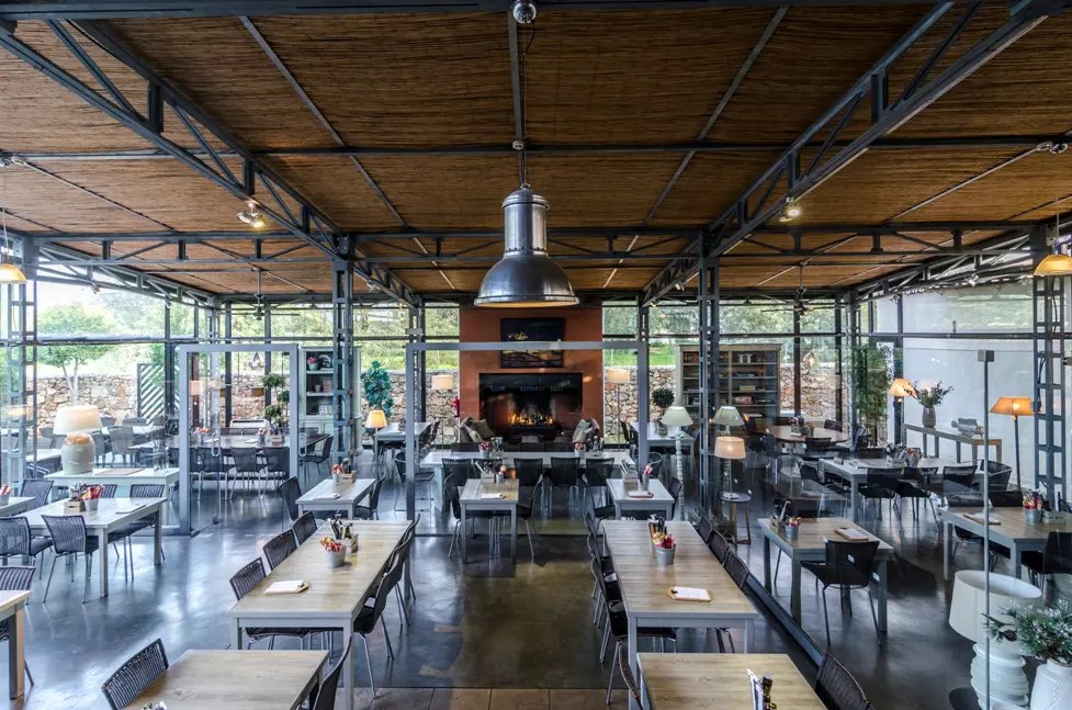 The dining room at Pizzeria Casavostra, Almancil — exposed steel and timber beams, long tables and a fireplace in the former Alquatro showroom
