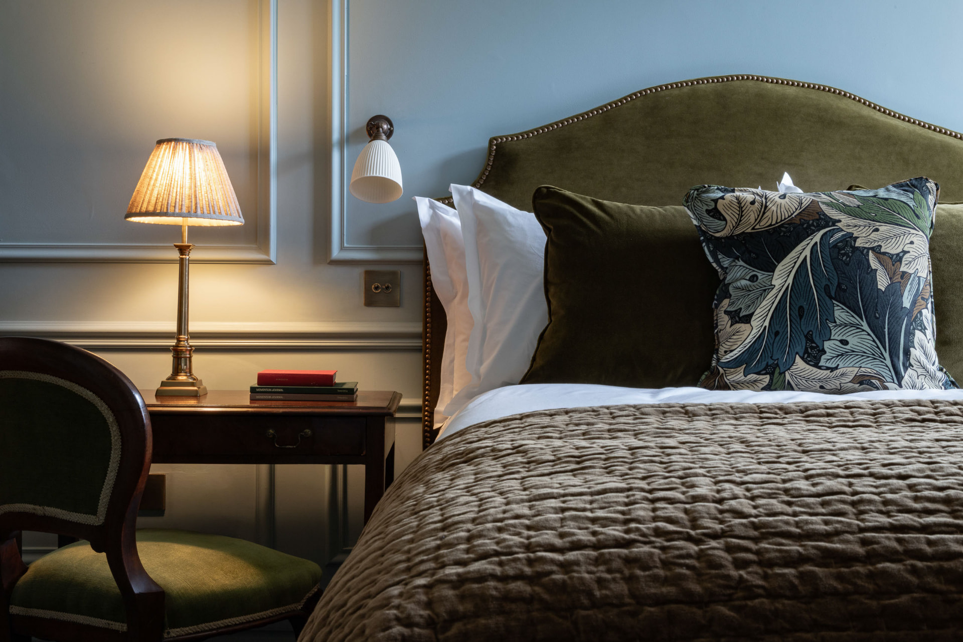 A bedroom at Dumbleton Hall — olive velvet headboard, patterned cushion and a lit bedside lamp
