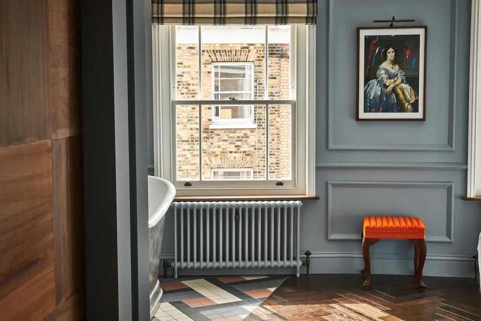 A blue panelled room with a sash window, an orange velvet stool and a framed portrait at The Lost Poet, Notting Hill