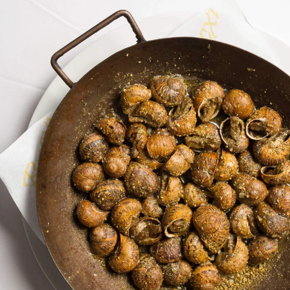Snails in a metal pan at El Racó del Cargol, seasoned and served on a white plate