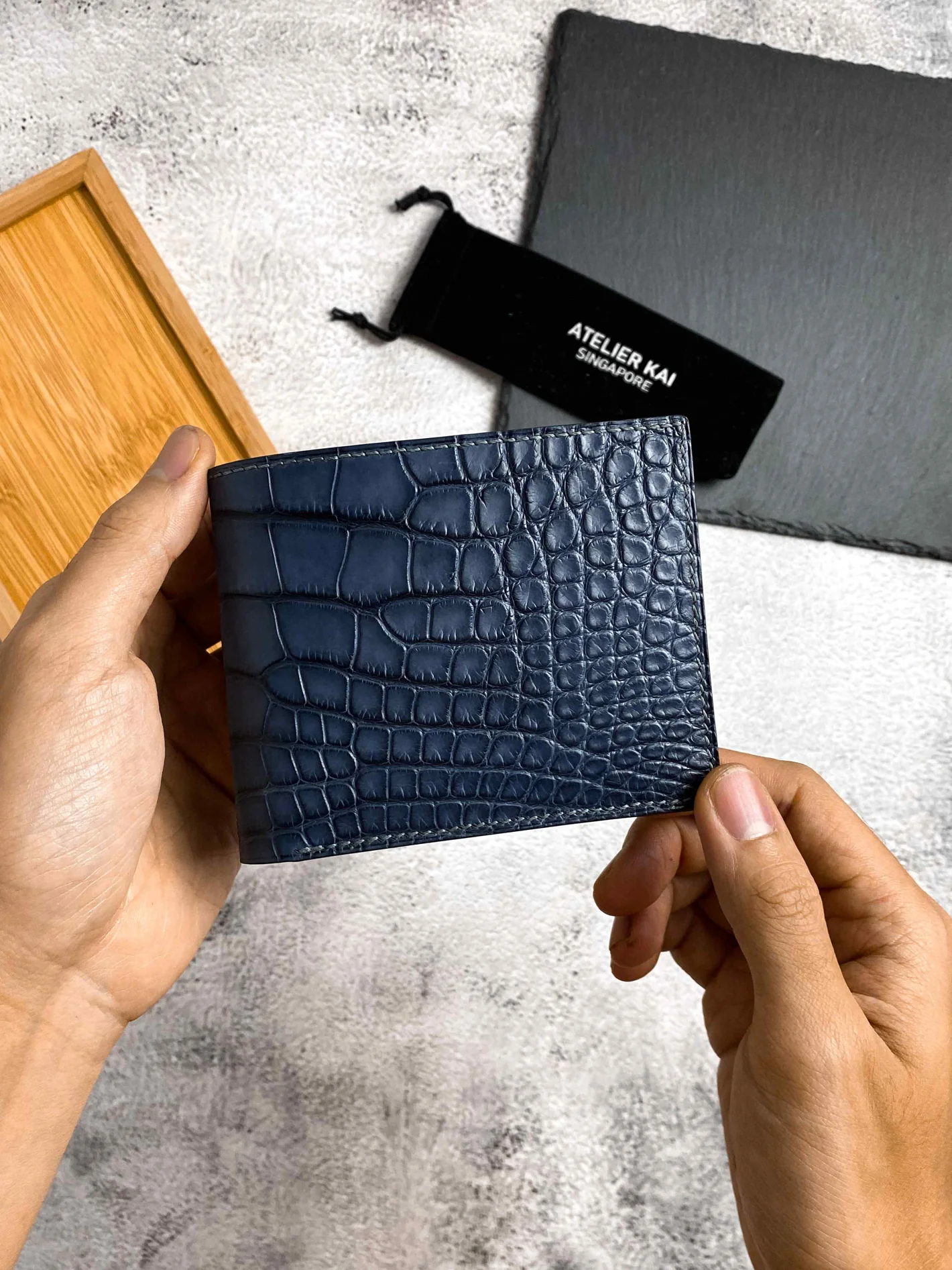 A navy crocodile-textured Atelier KAI wallet held up beside its packaging