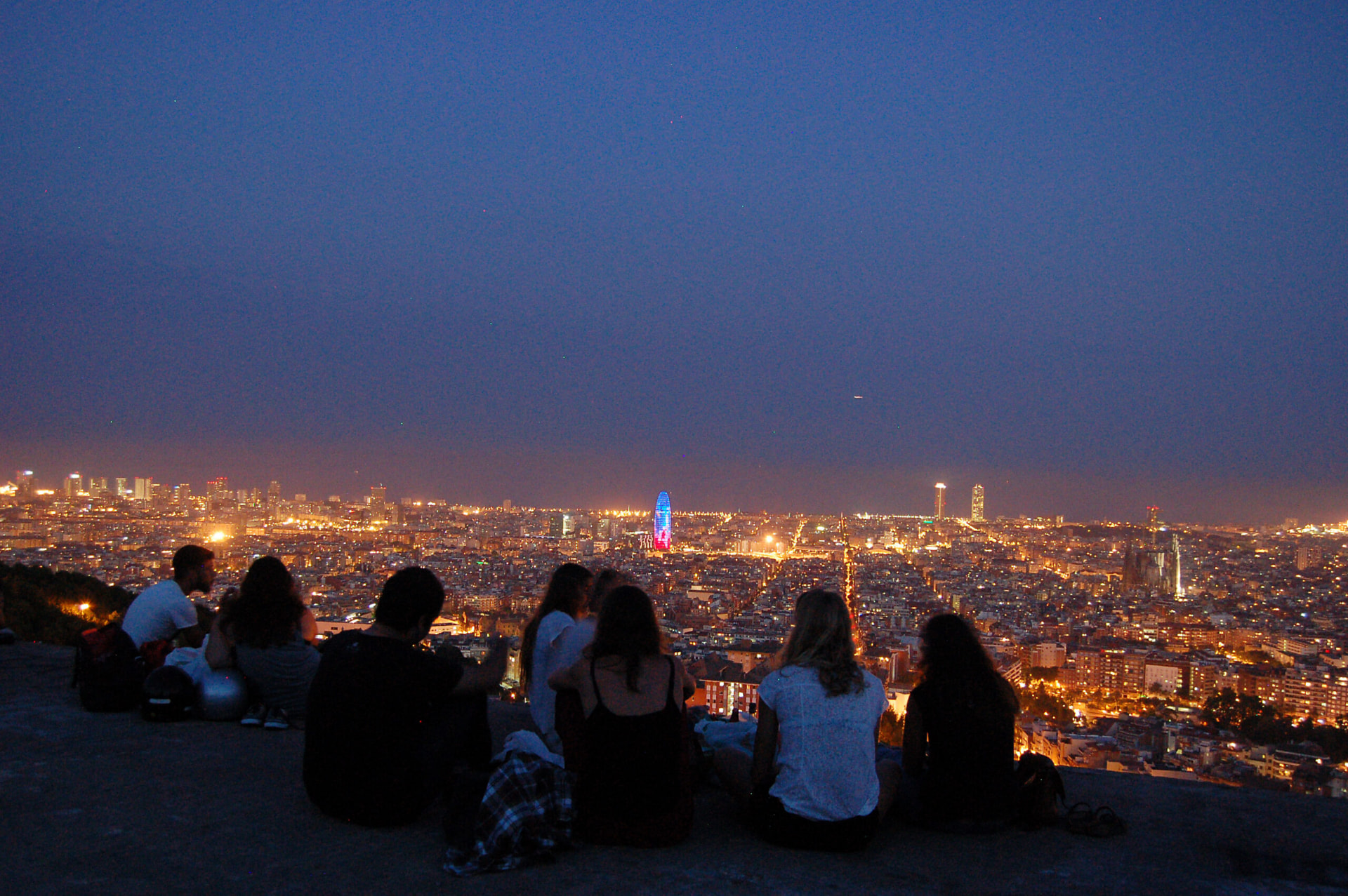 People sitting above Barcelona at dusk on the Turó de la Rovira, with the city lights and Torre Glòries below
