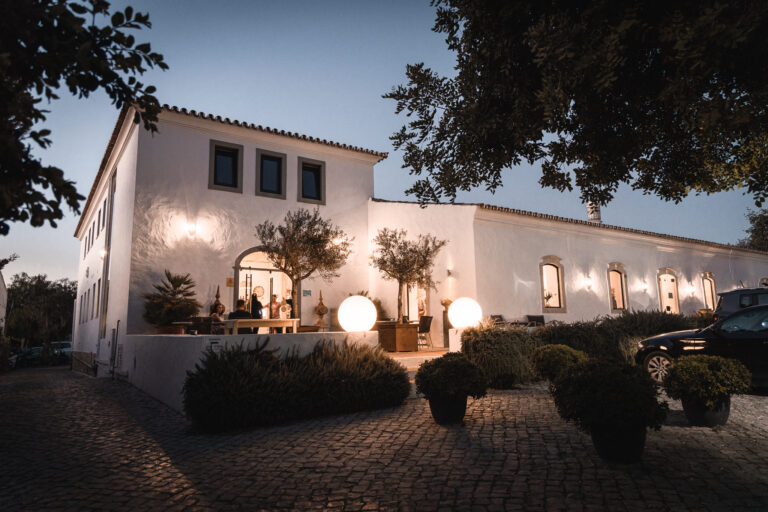 The white building of Pizzeria Casavostra at dusk, its terrace lit by glowing globe lamps