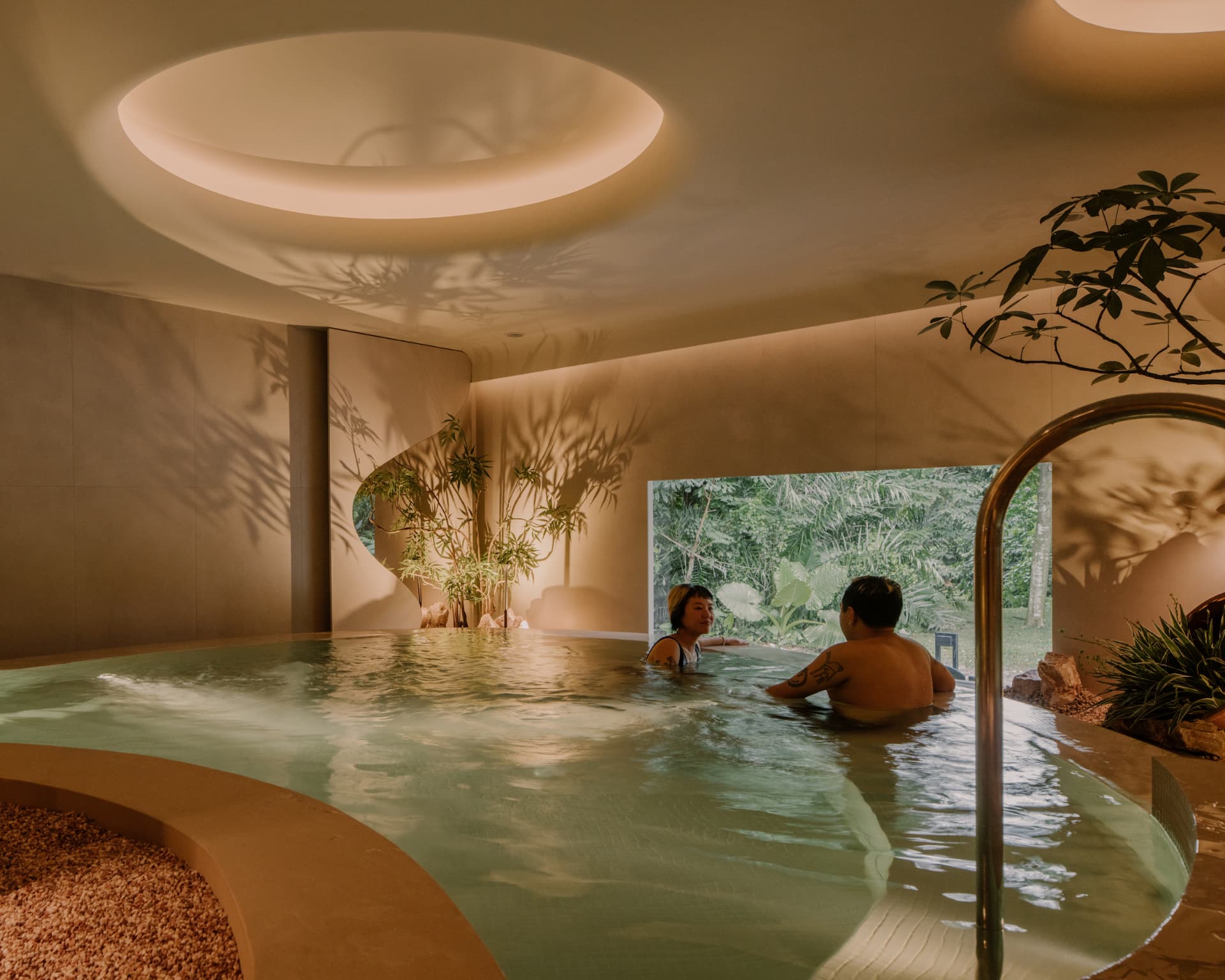 Two guests in a warm indoor pool at Nowhere Baths beneath a circular skylight