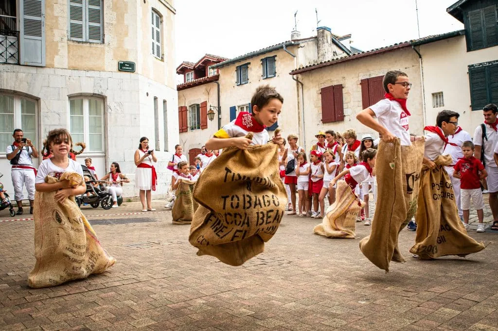 Children in white and red racing in hessian sacks through a Bayonne street during the Fêtes