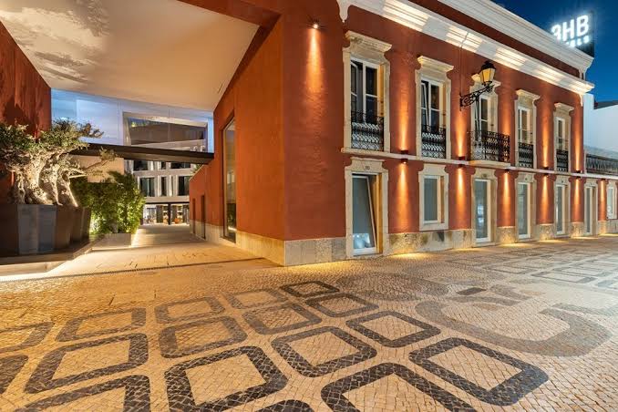 The red façade of 3HB Faro lit at dusk above a patterned Portuguese cobbled pavement