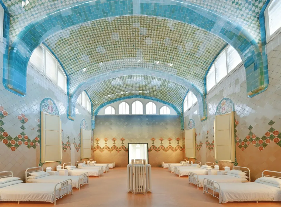 A restored Sant Pau hospital ward with white beds, tiled walls, floral ceramics and blue arched ceilings